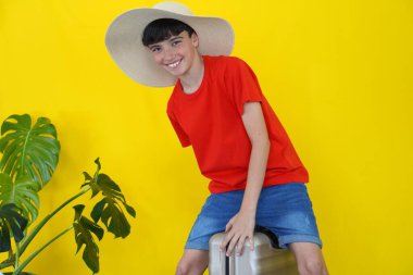 seyahat ve tatil konsepti. Şapkalı gülümseyen çocuk, elinde canavar yapraklarıyla bir bavulun üstünde oturuyor.