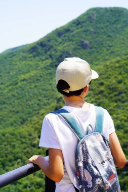 Dağları gözetleyen genç bir çocuğun yüzsüz görüntüsü. Bir yürüyüş turu sırasında bir kep ve sırt çantası takmış gözcülük yapıyor.