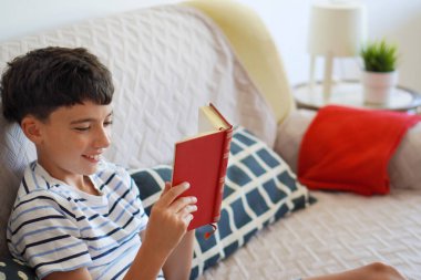 Çocuk kanepede kitap okurken gülümsüyor.