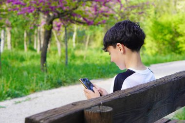 Çocuk sırtüstü oturmuş, bankta cep telefonuyla yazı yazıyor.