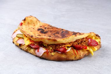 Füme jambon, baharatlı sosis, çedar peyniri, salatalık turşusu ve kızarmış soğanla doldurulmuş Rustik pizza sandviçi. Yanmış kabuk, açık gri taş zemin üzerindeki renkli dolgularla çelişiyor. Menüler ve reklamlar için mükemmel..