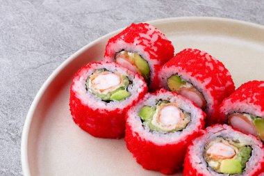 Parlak kırmızı tobiko ile kaplanmış karides tempura ve avokado ters yüz edilmiş, bej bir tabakla taş bir yüzey üzerinde servis edilir..