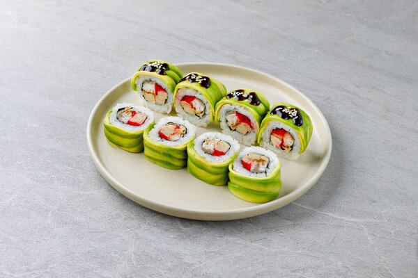 Avocado-wrapped sushi roll filled with crab stick and eel, teriyaki glaze and sesame on stone plate.