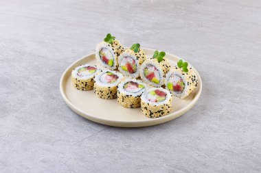 Taze ton balığı, taklit yengeç ve avokado içeren içi dışı suşi rulosu, bol miktarda siyah ve beyaz susam tohumlarıyla kaplanmış, mikroyeşilliklerle süslenmiş ve minimalist bej bir tabakta taş desenli bir zemin üzerinde servis ediliyor..