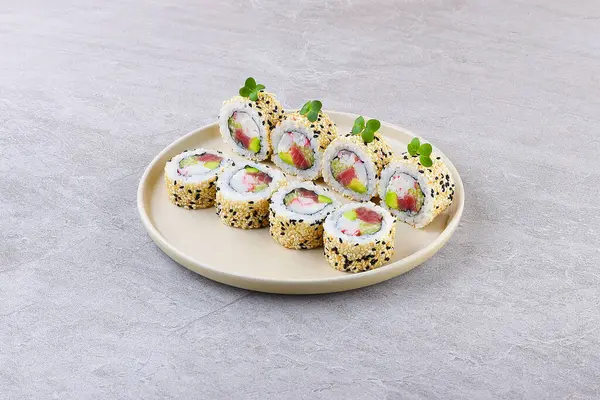 Taze ton balığı, taklit yengeç ve avokado içeren içi dışı suşi rulosu, bol miktarda siyah ve beyaz susam tohumlarıyla kaplanmış, mikroyeşilliklerle süslenmiş ve minimalist bej bir tabakta taş desenli bir zemin üzerinde servis ediliyor..