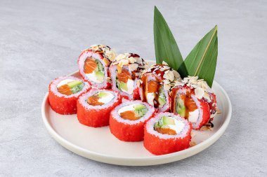 Kırmızı tobiko ile kaplanmış suşi ruloları ve taze somon, kremalı avokado ve keskin krem peynirle doldurulmuş, tatlı soya jölesiyle serpiştirilmiş ve bambu yaprağı desenli kavrulmuş badem dilimleri ile kaplanmış..