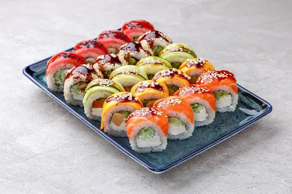 Somon, ton balığı, mango ve avokado dilimleriyle kaplı canlı gökkuşağı suşi ruloları, krem peynir ve salatalıkla doldurulmuş, yılan balığı sosu serpilmiş ve susam tohumlarıyla serpiştirilmiş..