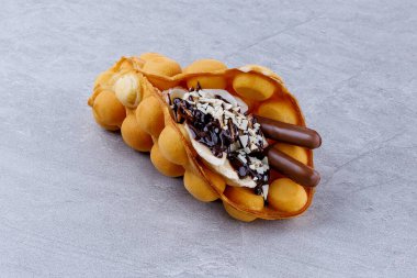 El yapımı bir koniye katlanmış altın yumurta waffle 'ı dilimlenmiş muz, krema, çikolata çubukları, badem taneleri ve zengin çikolata parçacıkları ile dolu, kırsal gri bir taş yüzeyde sunuluyor..