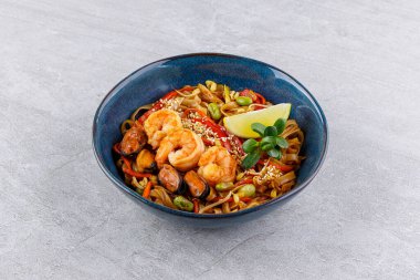 Dolgun karides, sulu midye, gevrek fasulye filizi ve edamame, parlak kırmızı çan biberi şeritleri ve susam tohumlarıyla süslenmiş canlı kızarmış pirinç eriştesi.