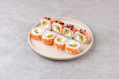 Avokado ile doldurulmuş, yumuşak karides dilimleri ve yılan balığı sosuyla kaplanmış, çıtır çıtır pirinç incileriyle süslenmiş, nötr bir arkaplana karşı minimalist bir seramik tabağa seramik bir suşi rulosu..