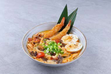 Kızarmış karides tempura, haşlanmış yumurta, mantar, domates, soğan ve erişteli otantik Japon eriştesi, gri arka planda seramik kasede servis ediliyor..