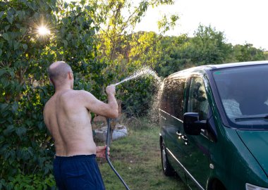 Adam yaz bahçesinde minibüsünü hortumla yıkıyor.