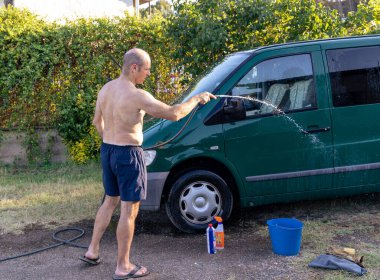 Adam bahçede minibüsünü birkaç temizlik ürünüyle yıkıyor.