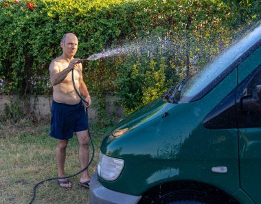Adam bahçede hortumla minibüsü yıkıyor.
