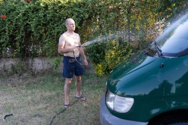 Genç adam bahçede suyla araba yıkıyor.