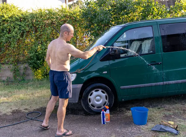 Adam bahçede minibüsünü birkaç temizlik ürünüyle yıkıyor.