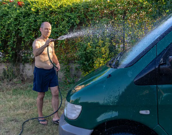 Adam bahçede hortumla minibüsü yıkıyor.