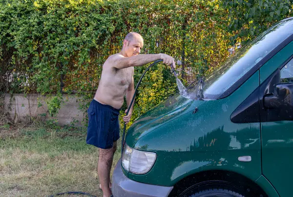 Bahçede hortumla araba yıkayan bir adam.