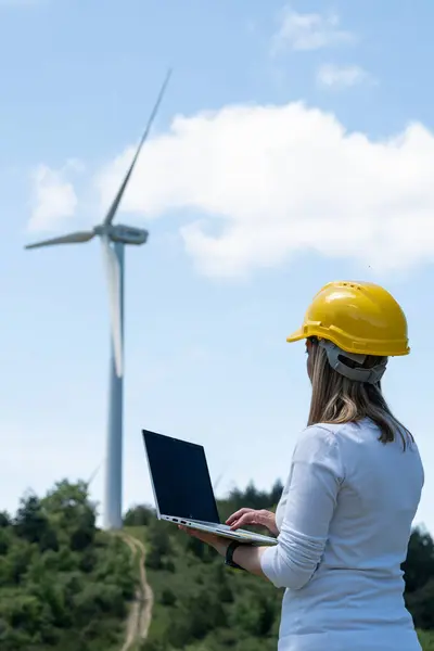 Mavi gökyüzünün arka planında dizüstü bilgisayarı ve rüzgar türbini olan kasklı kadın. Sürdürülebilir kalkınma ve enerji kavramı.