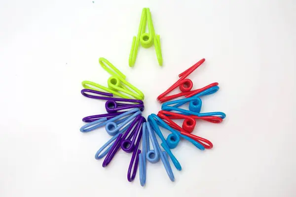 Renkli plastik mandallardan oluşan canlı bir tepeden tırnağa resim. Temiz beyaz bir arkaplanda dairesel bir desen oluşturulmuş. Simetrik düzen ve gökkuşağı renkleri, kavramlara uygun, görsel olarak çekici minimum bir kompozisyon oluşturur..