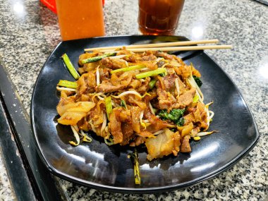 Lezzetli bir kızarmış Kwetiau eriştesi, sebze ve etle karıştırılmış, mükemmel pişirilmiş ve lezzetli bir yemek deneyimi için siyah bir tabakta sıcak servis edilmiş..