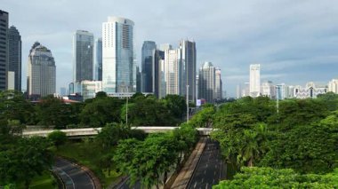 Yüksek binaları ve yeşil parkı olan bir şehir manzarası. Gökyüzü bulutlu ve şehir hareketli görünüyor.