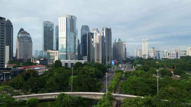 Yüksek binaları ve karayolu üzerinde köprüsü olan bir şehir. Gökyüzü bulutlu ve şehir hareketlilik içinde.