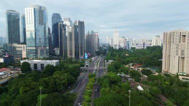 Yüksek binaları ve yeşil parkı olan bir şehir manzarası. Gökyüzü bulutlu ve şehir hareketli görünüyor.