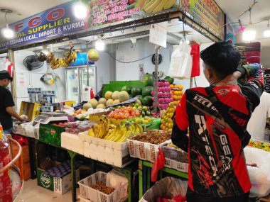 Kırmızı ve siyah gömlekli bir adam bir meyve standına bakıyor. Tribün muz, elma ve portakallarla dolu.