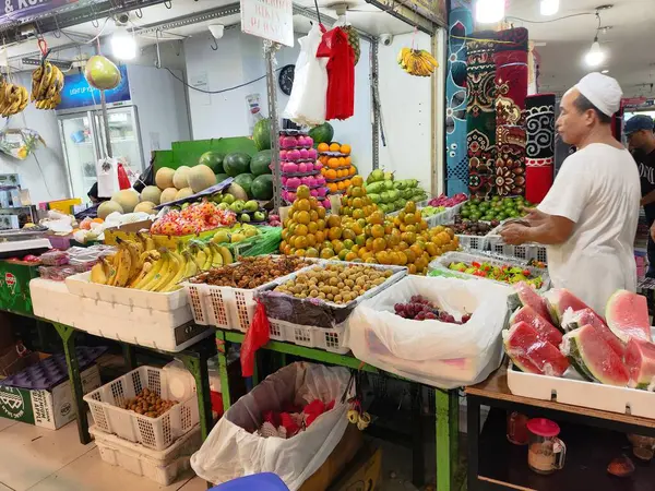 Jakarta, Endonezya - Nisan 12025: Kırmızı ve siyah gömlekli bir adam bir meyve standına bakıyor. Tribün muz, elma ve portakallarla dolu.