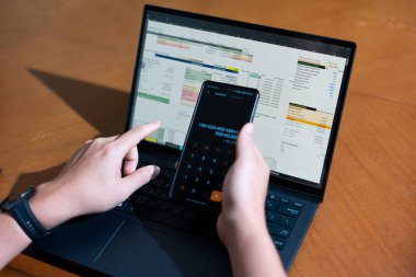 Bir kişi cep telefonu ve dizüstü bilgisayarı aynı anda kullanıyor. Cep telefonunda bir hesap makinesi var. Dizüstü bilgisayar ekranında çeşitli sayılar ve hesaplamalarla birlikte bir hesap tablosu gösteriliyor