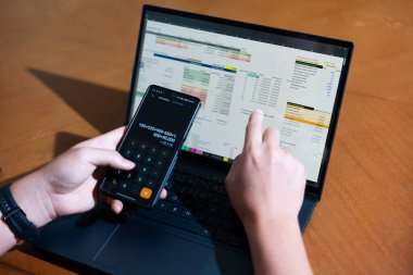 Bir kişi cep telefonu ve dizüstü bilgisayarı aynı anda kullanıyor. Cep telefonunda bir hesap makinesi var. Dizüstü bilgisayar ekranında çeşitli sayılar ve hesaplamalarla birlikte bir hesap tablosu gösteriliyor