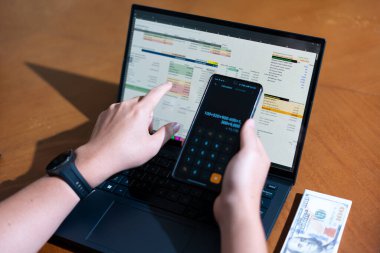 Bir kişi cep telefonu ve dizüstü bilgisayarı aynı anda kullanıyor. Cep telefonunda bir hesap makinesi var. Dizüstü bilgisayar ekranında çeşitli sayılar ve hesaplamalarla birlikte bir hesap tablosu gösteriliyor