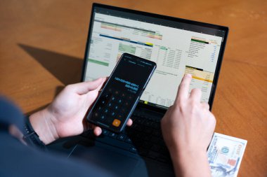 Bir kişi cep telefonu ve dizüstü bilgisayarı aynı anda kullanıyor. Cep telefonunda bir hesap makinesi var. Dizüstü bilgisayar ekranında çeşitli sayılar ve hesaplamalarla birlikte bir hesap tablosu gösteriliyor