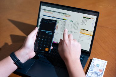 Bir kişi cep telefonu ve dizüstü bilgisayarı aynı anda kullanıyor. Cep telefonunda bir hesap makinesi var. Dizüstü bilgisayar ekranında çeşitli sayılar ve hesaplamalarla birlikte bir hesap tablosu gösteriliyor