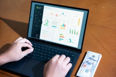 Bir kişi yeşil ekranlı bir dizüstü bilgisayarda yazıyor. Laptop üzerinde grafik olan bir sayfayı açıyor. Laptopun yanındaki masada 100 dolarlık banknotlar var.