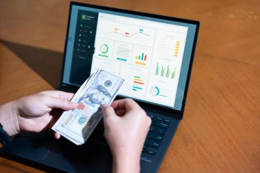 Bir kişi finansal sisteme para girmek için dizüstü bilgisayar kullanıyor. Kişi bir yığın banknot tutuyor ve dizüstü bilgisayarda paranın giriş yaptığı bir grafik var. Sahne ciddi ve odaklı.