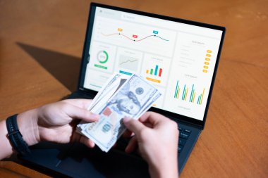 Bir insan 100 dolarlık banknotları dizüstü bilgisayarın önünde tutuyor. Laptop ekranında para yığınının altında bir çizgi var.