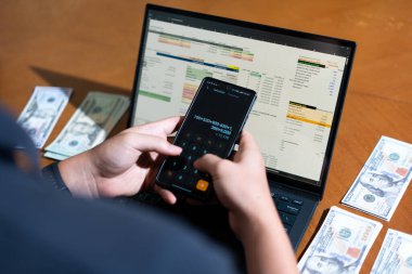 Bir kişi toplam para miktarını hesaplamak için bir dizüstü bilgisayar ve bir cep telefonu kullanıyor. Dizüstü bilgisayar ekranında çeşitli sayılar ve hesaplamalarla birlikte bir hesap tablosu gösteriliyor