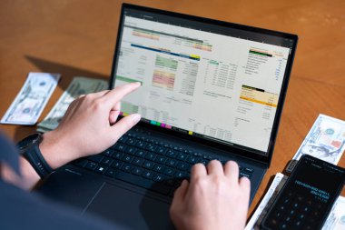 Bir kişi hesap tablosuna bakmak için dizüstü bilgisayar kullanıyor. Laptop hesap makinesi ve cep telefonu olan bir masada.