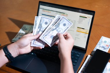 Bir kişi para saymak için dizüstü bilgisayar kullanıyor. Adamın elinde bir yığın banknot ve hesap makinesi var.