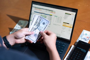 Bir kişi para saymak için dizüstü bilgisayar kullanıyor. Adamın elinde bir yığın banknot ve hesap makinesi var.