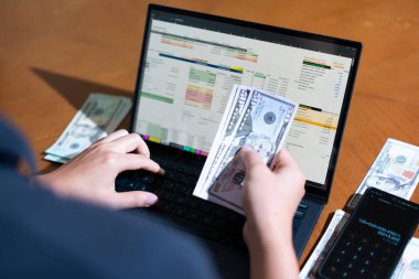 Bir kişi para saymak için dizüstü bilgisayar kullanıyor. Adamın elinde bir yığın banknot ve hesap makinesi var.