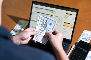 Bir kişi para saymak için dizüstü bilgisayar kullanıyor. Adamın elinde bir yığın banknot ve hesap makinesi var.