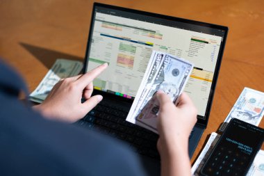 Bir kişi para saymak için dizüstü bilgisayar kullanıyor. Adamın elinde bir yığın banknot ve hesap makinesi var.