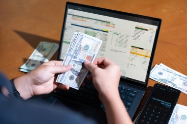 Bir kişi para saymak için dizüstü bilgisayar kullanıyor. Adamın elinde bir yığın banknot ve hesap makinesi var.
