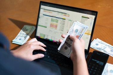 Bir kişi para saymak için dizüstü bilgisayar kullanıyor. Adamın elinde bir yığın banknot ve hesap makinesi var.