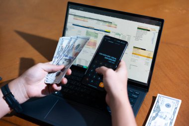 Bir kişi para saymak için dizüstü bilgisayar kullanıyor. Adamın elinde bir yığın banknot ve hesap makinesi var.