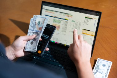 Bir kişi sahip olduğu para miktarını hesaplamak için bir dizüstü bilgisayar kullanıyor. Yakınlarda bir hesap makineleri ve cep telefonları var.