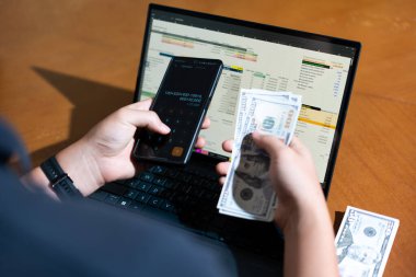 Bir kişi işlem yapmak için dizüstü bilgisayar ve cep telefonu kullanıyor. Laptopun bir hesap makinesi uygulaması açık ve cep telefonunda bir mesaj var. Adamın elinde yığınla para var.
